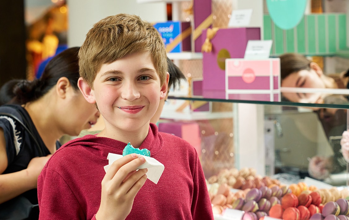 A smiling boy eating a Birthday Cake macaron. Behind him is a Woops! Kiosk full of assorted macarons and macaron boxes.