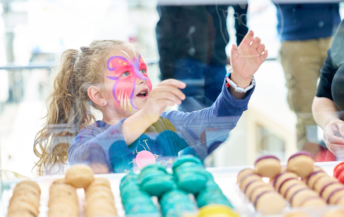 A little girl holding her arms up, in front of her are numerous assorted macarons.