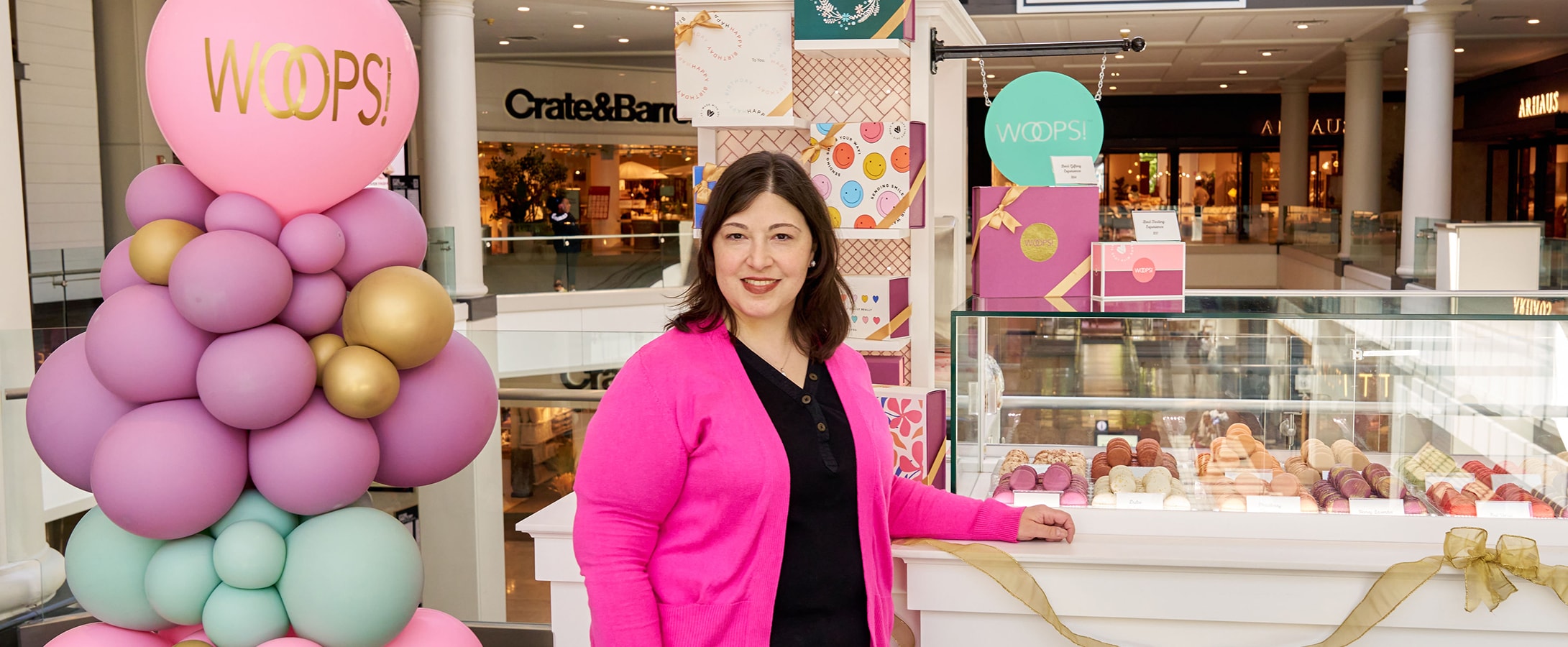 A smiling woman with a pink sweater standing in front of a Woops! Macarons & Gifts kiosk. To the left are some colorful balloons.