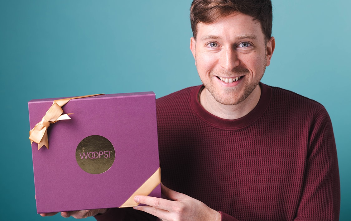 Smiling man with a red sweater holding a purple Woops! Macaron box. 