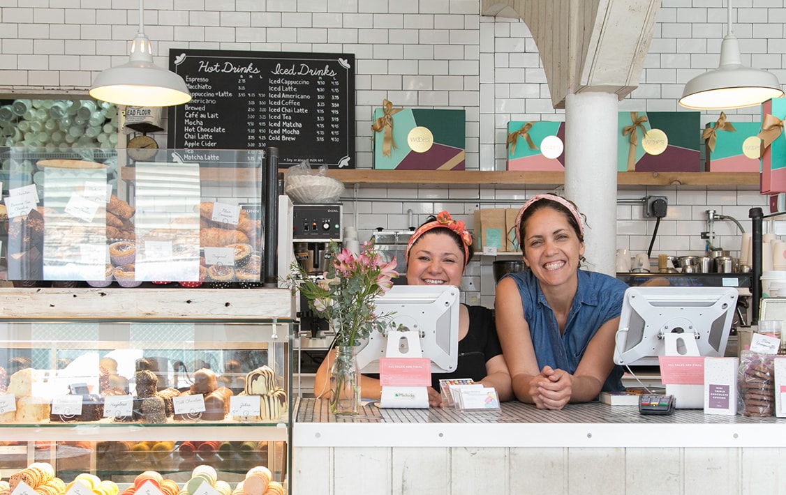 Two smiling women behind a counter full of assorted macaron boxes, macarons, and desserts.