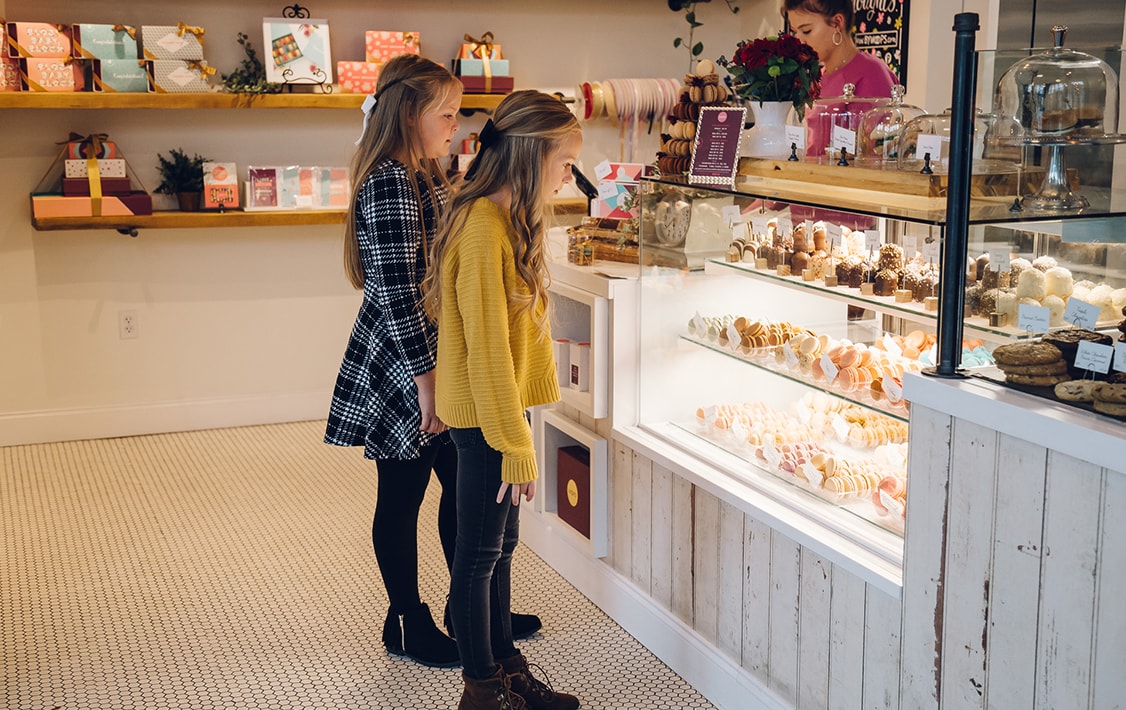 Two girls looking at a display of Woops! French macarons. Surrounding them are assorted macaron boxes and desserts.