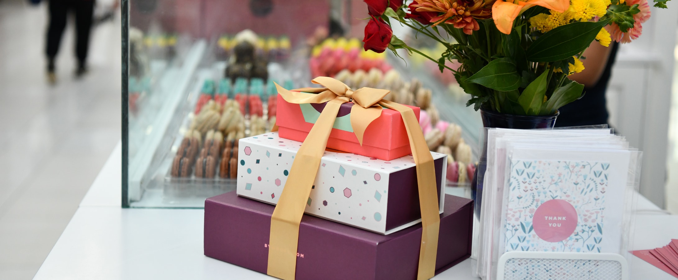 A Woops! Macarons & Gifts kiosk counter is full of assorted macarons boxes, greeting cards, a display of macarons, and flowers.