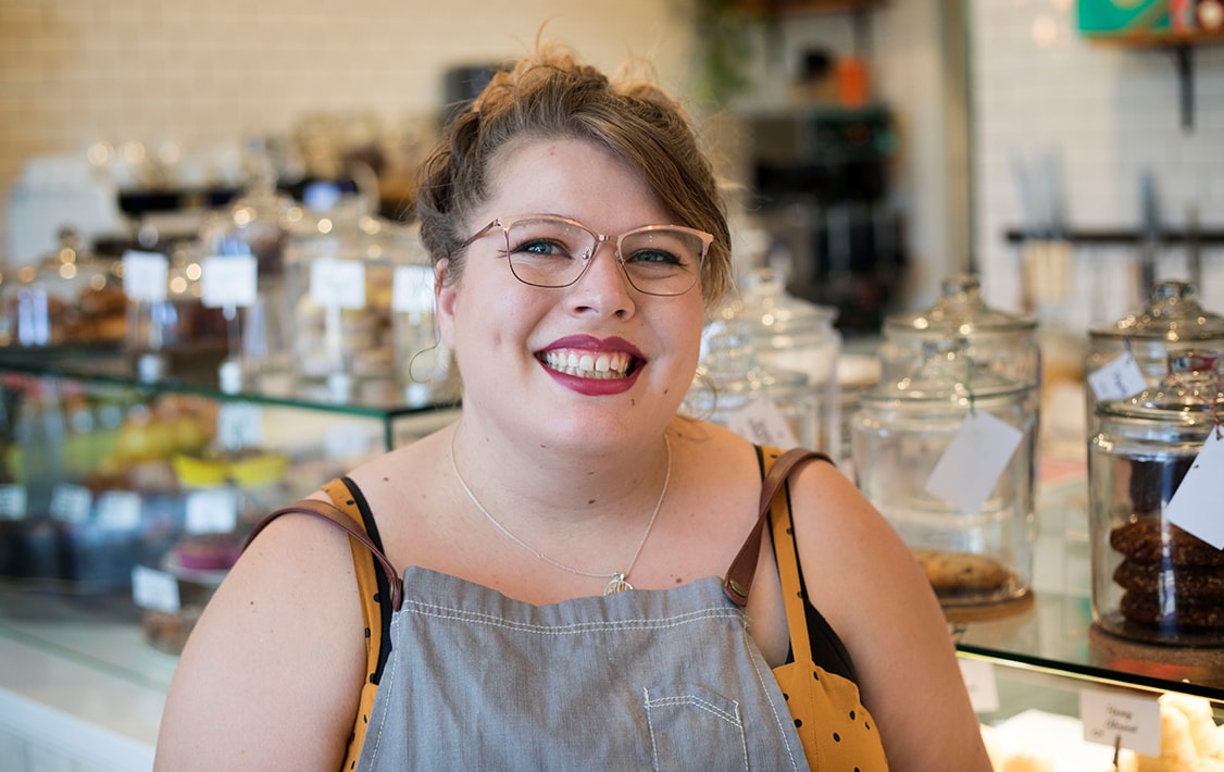 A smiling woman in front of a Woops! Bakeshop counter full of assorted macarons and cookies.