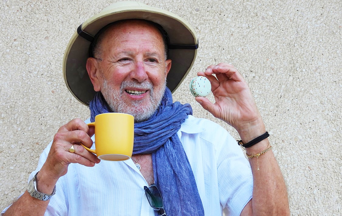 A smiling man holding a macaron in one hand and a coffee cup in the other.