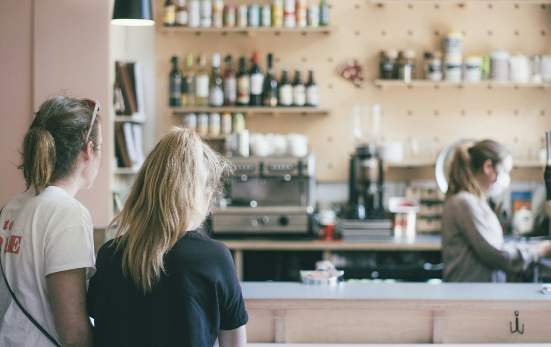 Two customers standing at a café counter watching a barista prepare drinks, with shelves of bottles and jars in the background.