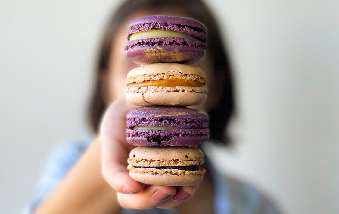A woman holding 4 Woops! macarons in line, purple and beige. 
