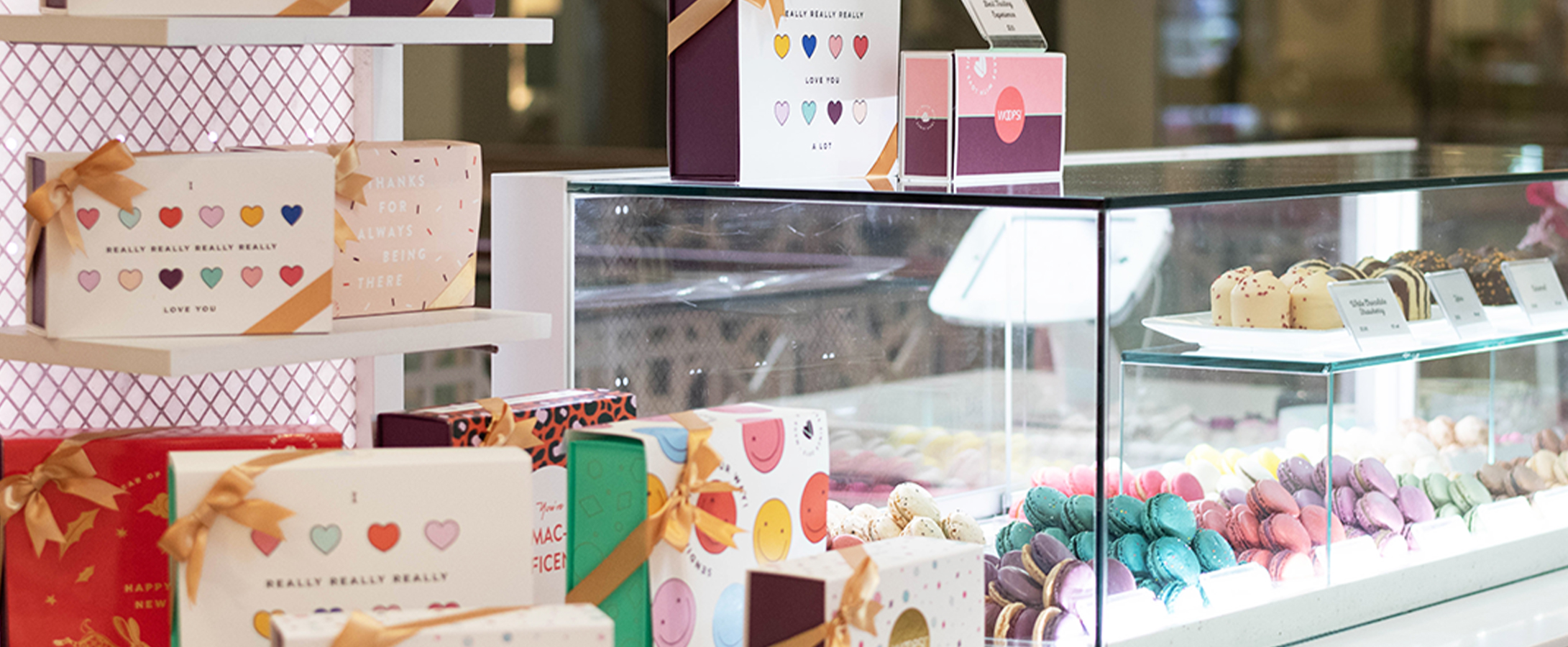 Woops! macaron display case filled with colorful macarons, surrounded by gift boxes with gold ribbons and playful designs inside a bright, elegant shop.