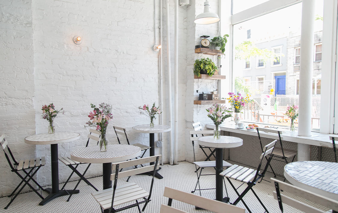 Bright, minimalist café interior with white brick walls, small round mosaic tables, folding chairs, and fresh flower arrangements by a large window letting in natural light. 