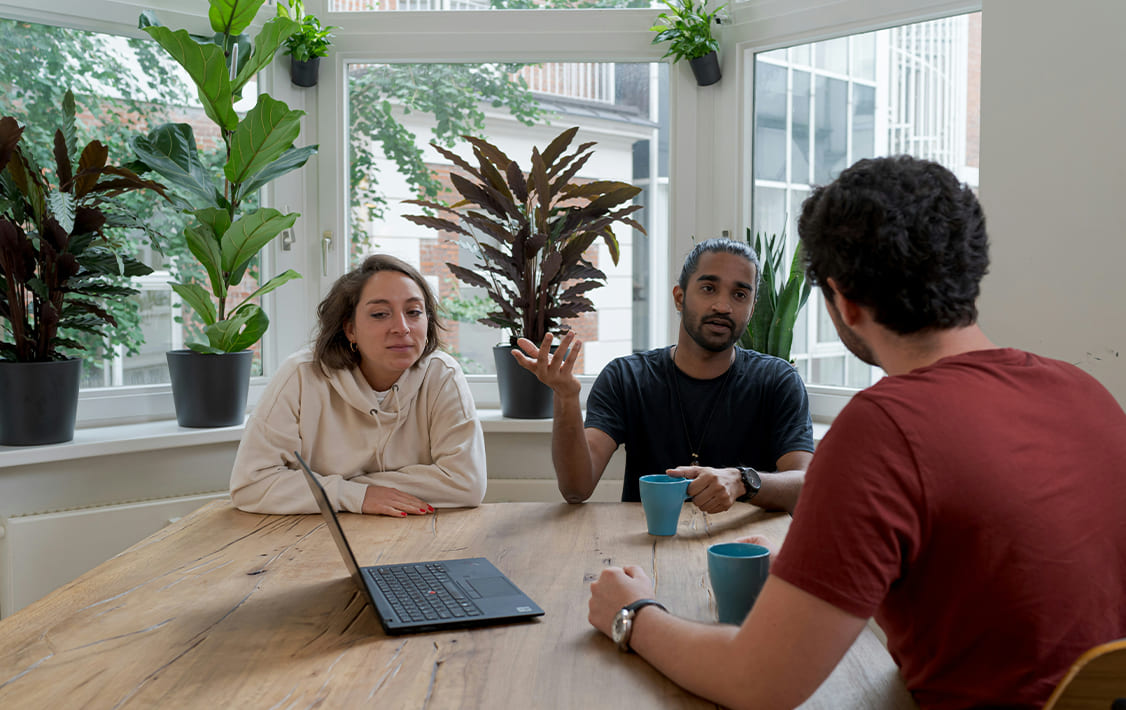 Three colleagues having a meeting around a wooden table with a laptop and coffee mugs, seated in a bright room with large windows and potted plants.