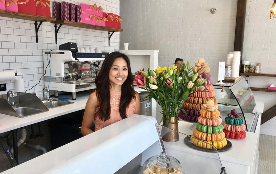 Smiling woman standing behind the counter of a bright macaron shop, with colorful macaron towers, fresh flowers, and pink gift boxes displayed against white subway tile walls.
