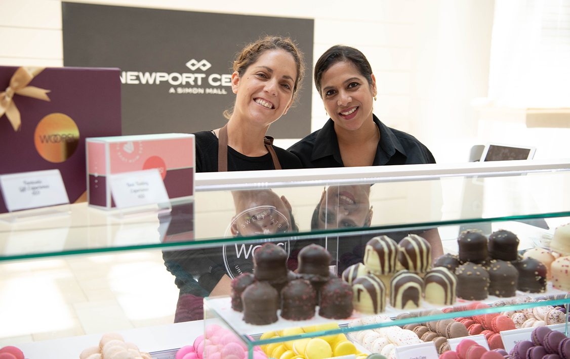 Two Woops! employees smile behind a macaron display at a Newport Centre mall kiosk.