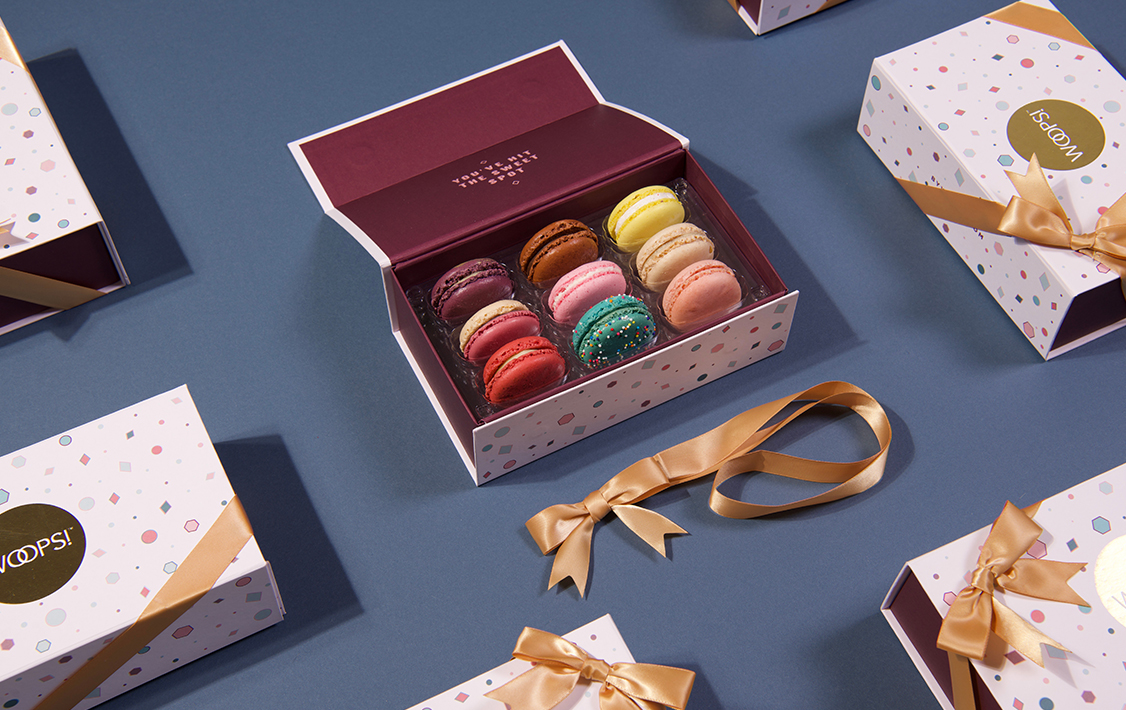 Open Woops! macaron box filled with colorful macarons on a blue surface, surrounded by closed gift boxes wrapped with gold ribbons.