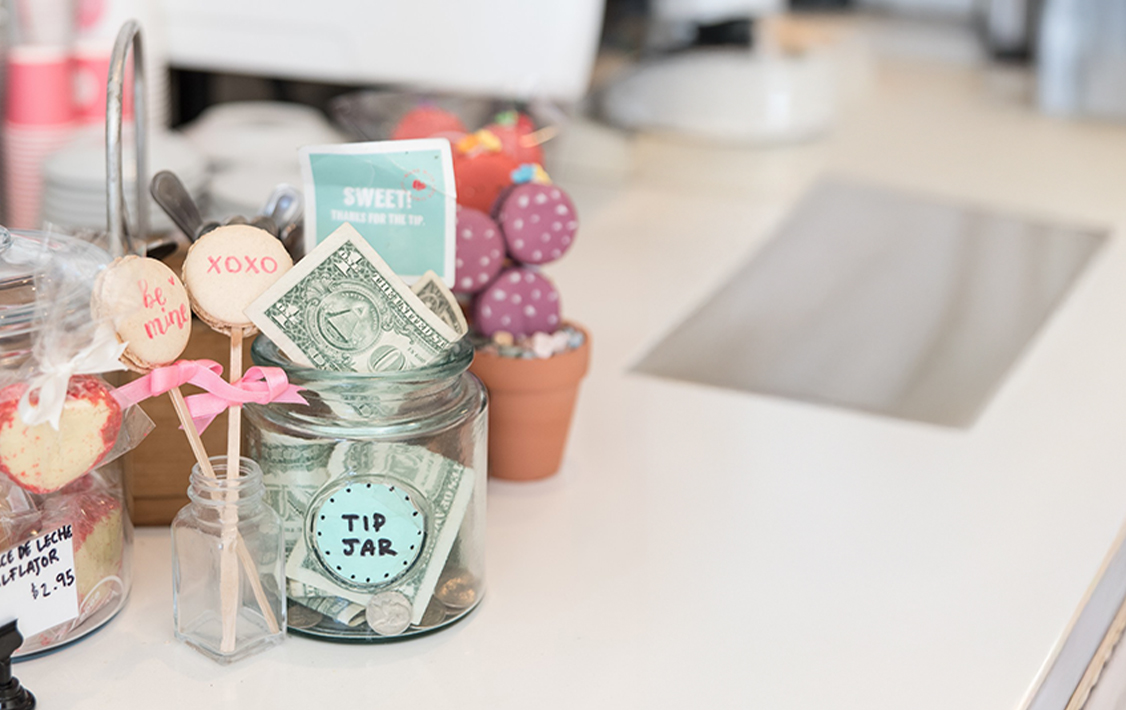 A café tip jar filled with dollar bills and coins, decorated with macaron pops labeled “be mine” and “xoxo,” surrounded by sweets and small display jars on the counter.