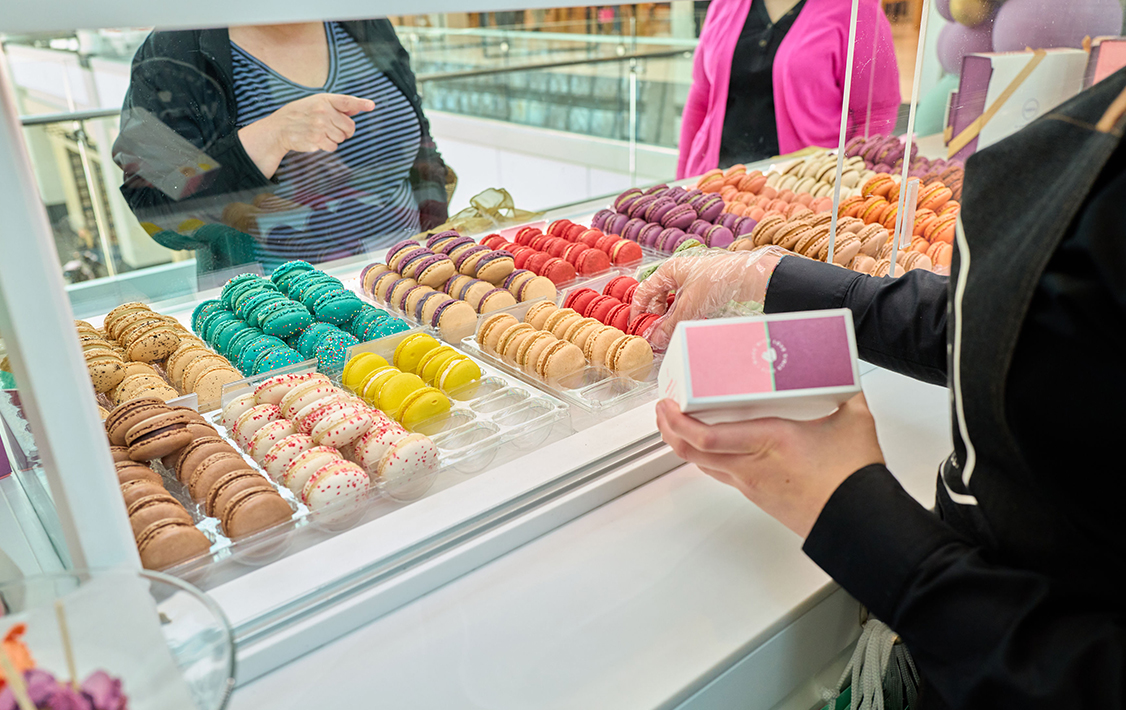 A Woops! employee packages macarons behind a glass display filled with colorful rows of macarons as customers point and choose their flavors.