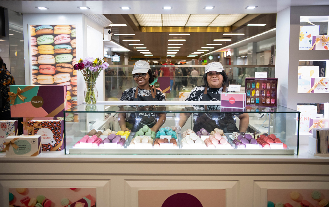 Two Woops! employees smile behind a macaron display case at a brightly lit kiosk, surrounded by colorful gift boxes and trays of assorted macarons.
